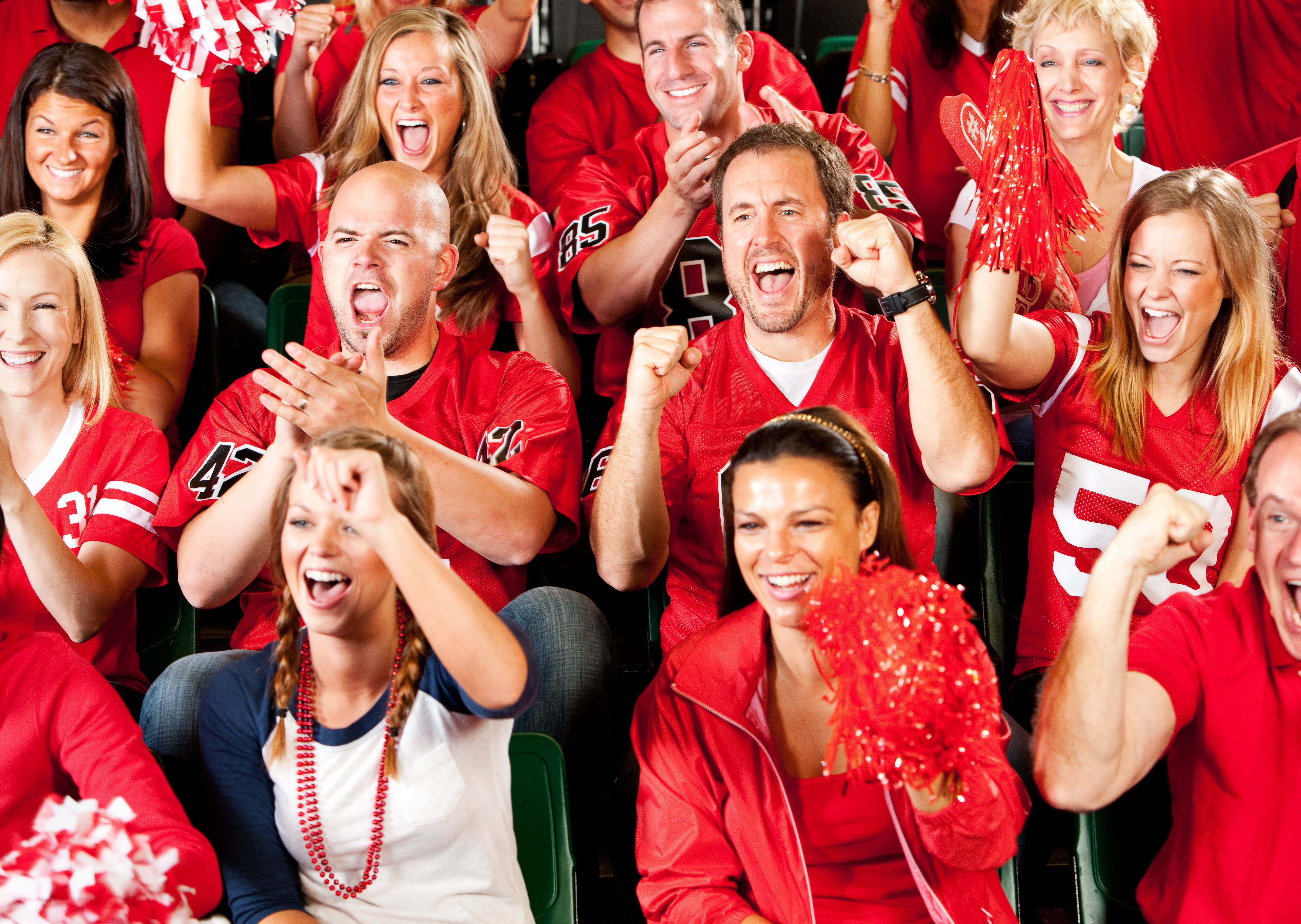 A crowd of fans in red team jerseys on their feet in the stands, cheering, clapping and waving pom-poms